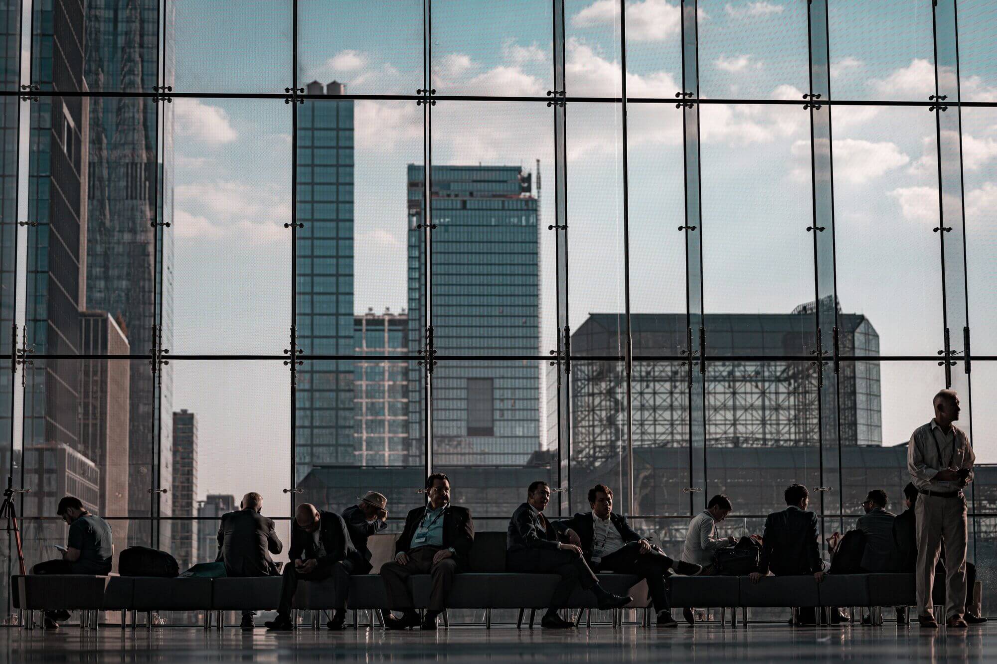 Business travelers with event badges await Javits Center transportation by a glass wall over the Manhattan skyline.