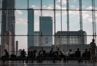 Business travelers with event badges await Javits Center transportation by a glass wall over the Manhattan skyline.